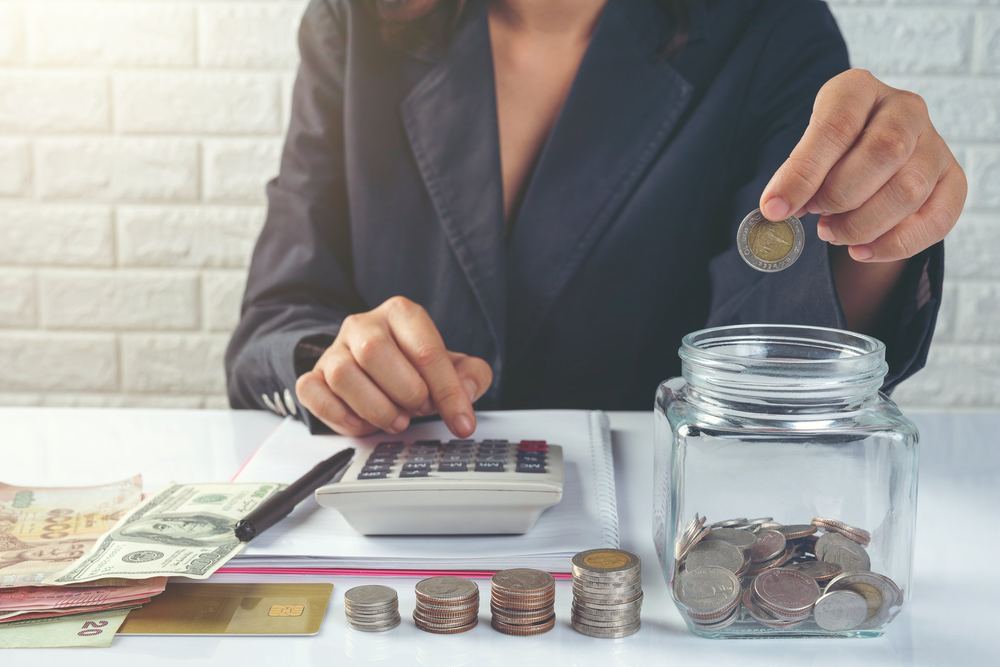 A hand dropping a coin into a jar beside a calculator and cash